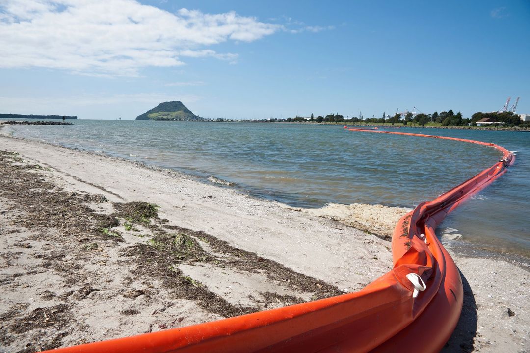 Sotto un cielo azzurro brillante, una barriera galleggiante arancione per il contenimento del petrolio si estendeva sulle acque costiere verso una montagna lontana.