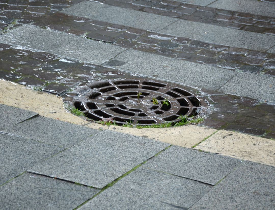 Una griglia di scolo circolare in ferro, incastonata in un percorso pedonale pavimentato e circondata da piastrelle di pietra grigio chiaro.