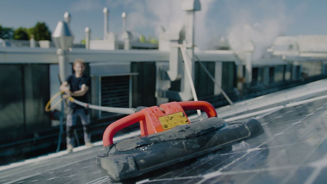 Una persona pulisce i pannelli solari su un tetto utilizzando un attrezzo manuale con manico rosso e spazzola nera.