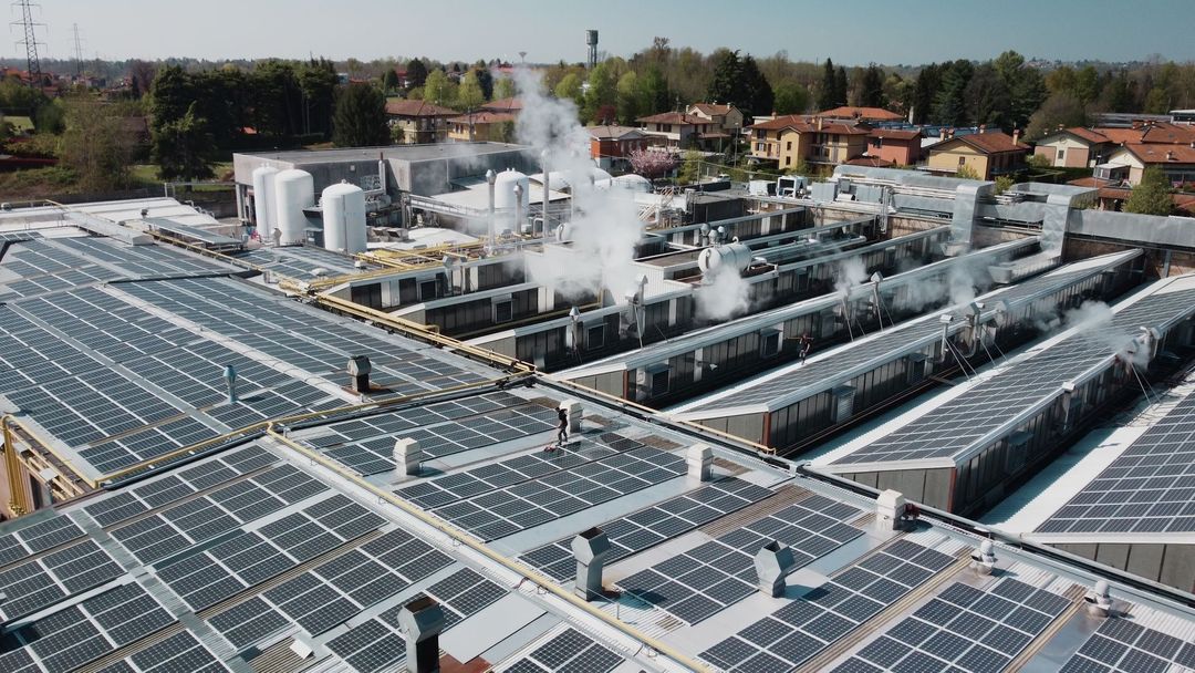 Il tetto di un edificio industriale ricoperto di pannelli solari, con vapore che fuoriesce dalle prese d'aria tra i pannelli, vicino ad alcune abitazioni.