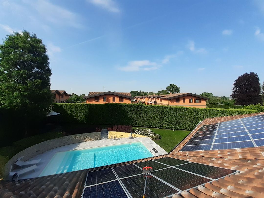 Una scena all'aperto illuminata dal sole che mostra una piscina in un giardino e un tetto ricoperto di pannelli solari, con delle case sullo sfondo.