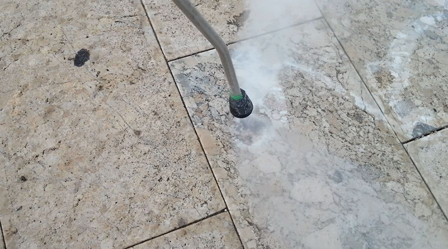 L'ugello di un'idropulitrice spruzza acqua su un patio di piastrelle di pietra sporche, rivelando una sezione sottostante pulita e più chiara.