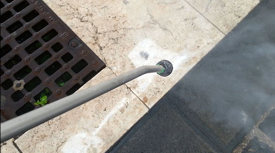 La lancia di un'idropulitrice spruzza un getto d'acqua ad alta pressione su una pavimentazione in pietra chiara vicino a una griglia di scarico metallica.