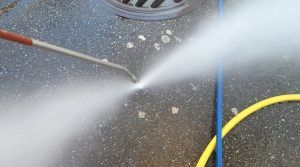 L'ugello di un'idropulitrice spruzza un potente getto d'acqua su una superficie di cemento vicino a un tubo giallo.