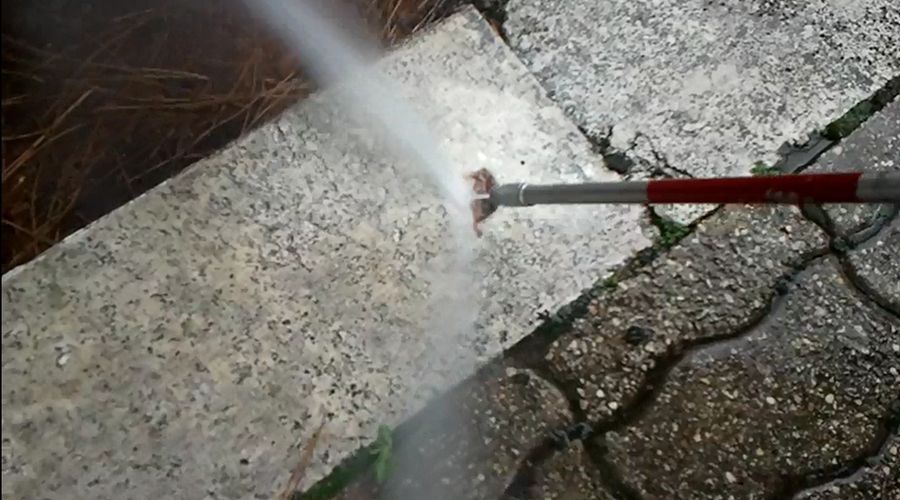 La lancia di un'idropulitrice spruzza acqua su una superficie pavimentata sporca, pulendo una sezione chiara del cemento.