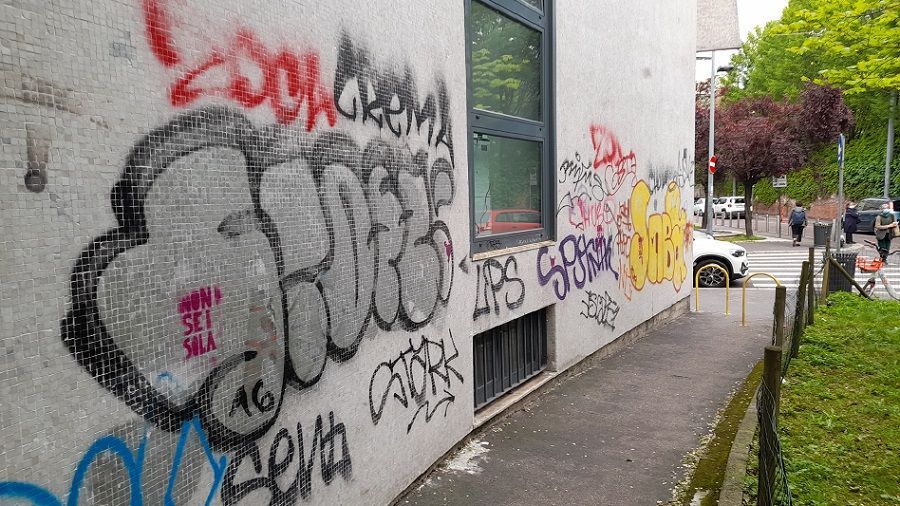 Un muro di cemento ricoperto di graffiti colorati e scritte in stile fumetto, adiacente a un marciapiede e a uno spazio verde.