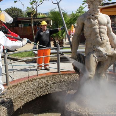 Operai con indumenti protettivi utilizzano un tubo ad alta pressione per pulire una grande statua di pietra in una fontana all'aperto.