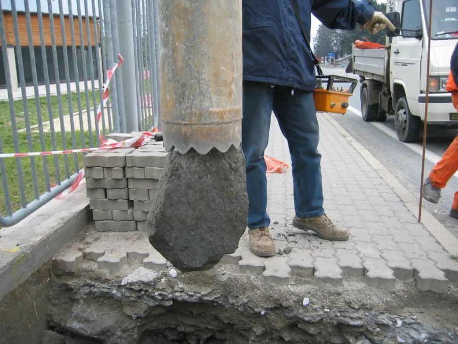 Un operaio edile tiene in mano uno strumento di carotaggio specializzato sopra un foro in un marciapiede asfaltato, vicino a un camion e a una recinzione metallica.