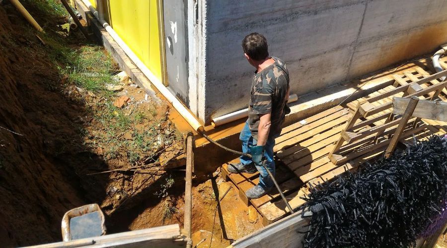 Una persona con una camicia a fantasia si trova su una piattaforma di legno accanto a una fondazione in cemento e a uno scavo di terra.
