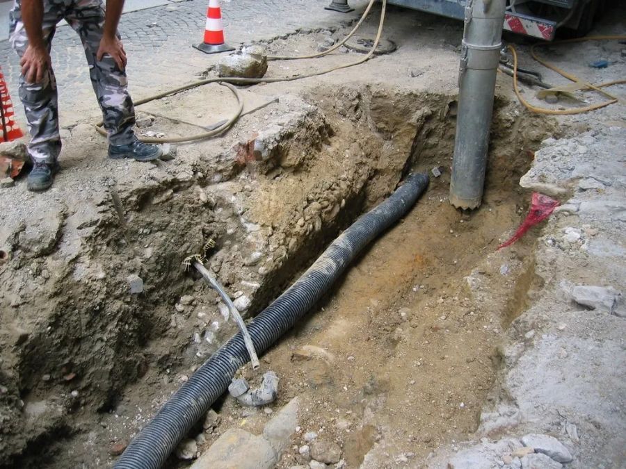 In un cantiere edile, un operaio si trova vicino a una trincea contenente un tubo di drenaggio nero e una colonna verticale di cemento.