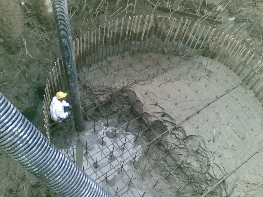 Un operaio con un elmetto giallo versa del calcestruzzo in una fossa circolare per le fondamenta, rinforzata con barre d'armatura e paletti di legno.