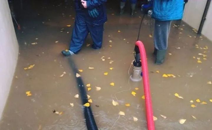 Due persone con indumenti impermeabili utilizzano una pompa per drenare l'acqua allagata da una stanza cosparsa di foglie gialle cadute.