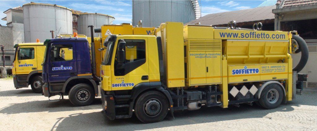 Tre camion commerciali, due gialli e uno blu scuro, sono parcheggiati in un piazzale ghiaioso vicino a dei silos industriali.