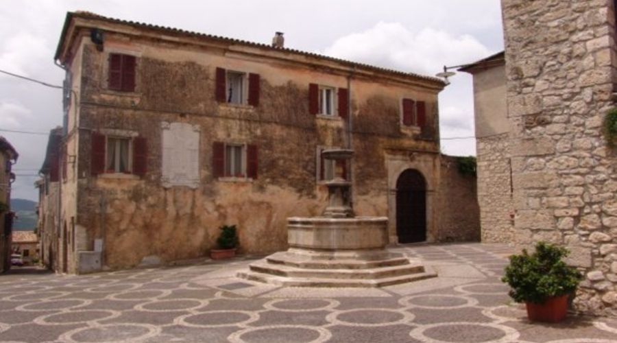 Una fontana in pietra si erge in una piazza del villaggio pavimentata con motivi circolari, fiancheggiata da storici edifici in pietra dallo stile rustico.