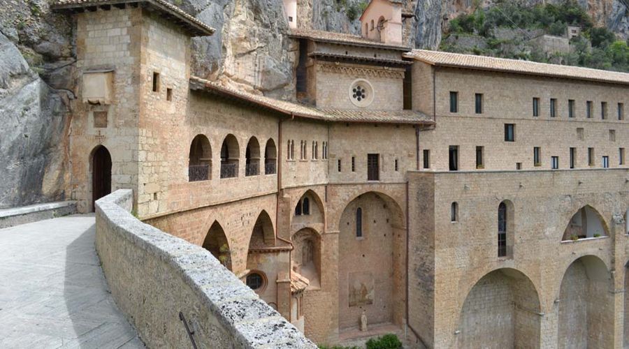 Monastero in pietra incastonato nella parete rocciosa di una scogliera, caratterizzato da passaggi ad arco e architettura in pietra, in Italia.