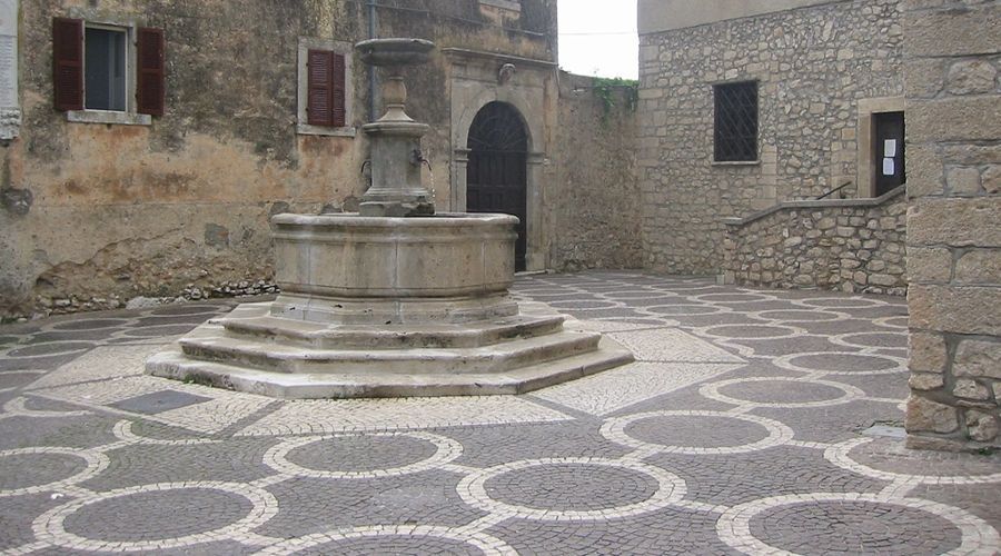 Al centro di un cortile lastricato si trova una fontana in pietra con un motivo geometrico di cerchi bianchi e grigi.