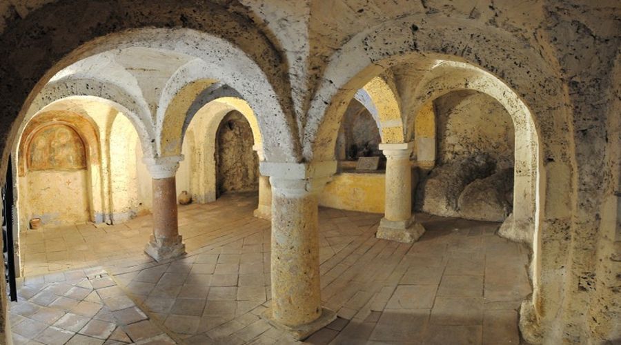Una cripta o cantina storica in pietra caratterizzata da soffitti a volta sorretti da robuste colonne e pareti dalla texture grezza.