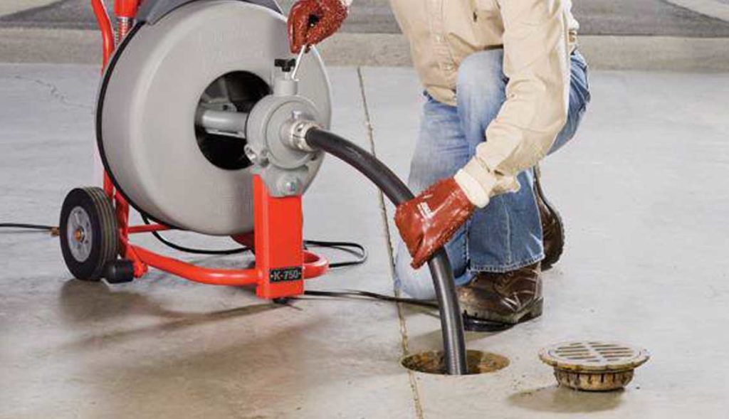 Un tecnico con i guanti inserisce una sonda motorizzata per scarichi in uno scarico a pavimento, utilizzando un'idraulica elettrica di colore rosso e grigio.