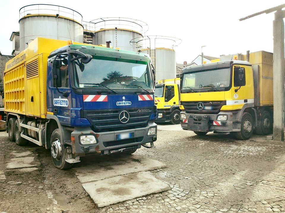 Tre grossi camion industriali parcheggiati in un piazzale ghiaioso accanto ad alti silos di stoccaggio.