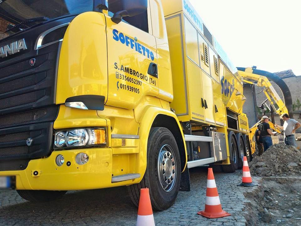 Un camion escavatore a risucchio di colore giallo brillante è parcheggiato su una strada acciottolata, con degli operai in piedi accanto a un cantiere di scavo.
