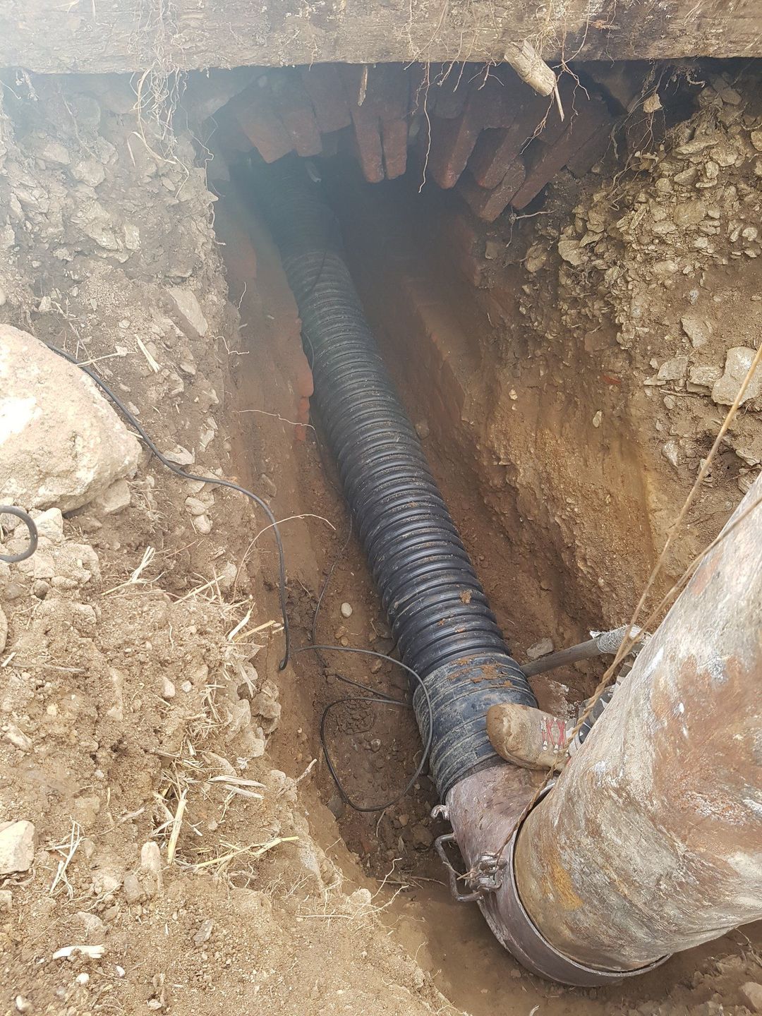Un tubo di plastica nera e corrugata è collegato a un tubo metallico più grande all'interno di una trincea riempita di terra, sotto una struttura in mattoni.
