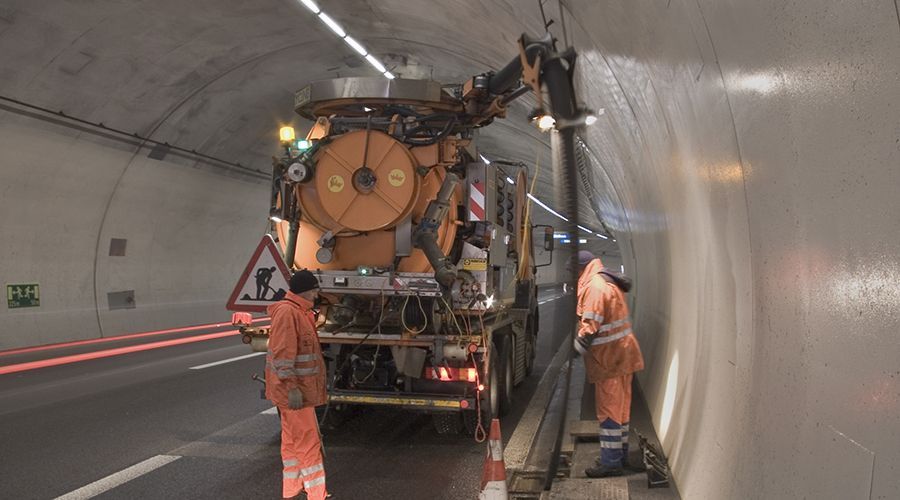 Operai con indumenti arancioni ad alta visibilità eseguono lavori di manutenzione all'interno di una galleria utilizzando un camion con braccio elevatore.