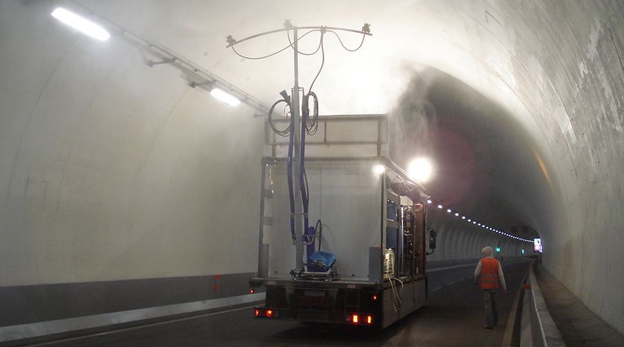 Un camion dotato di un sistema di pulizia sopraelevato nebulizza acqua all'interno di un tunnel ad arco ben illuminato, mentre un operaio cammina nelle vicinanze.