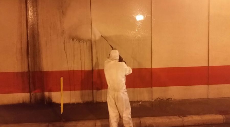 Una persona con indosso una tuta protettiva bianca utilizza un'idropulitrice per pulire una parete bianca con una striscia rossa orizzontale.