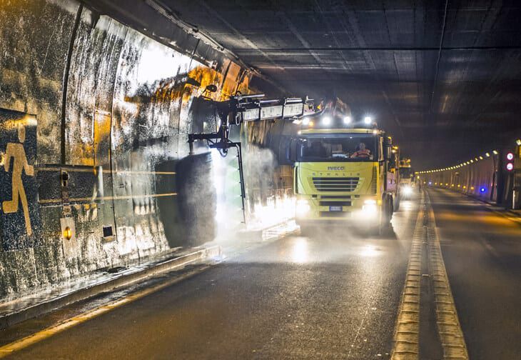 Un camion per le pulizie giallo utilizza un braccio robotico per spruzzare e strofinare la parete interna di una galleria buia.