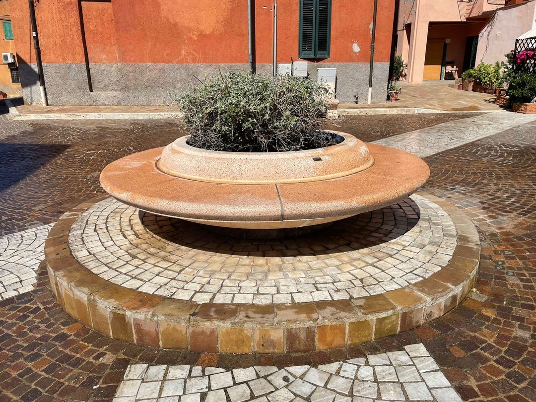 Al centro di un cortile lastricato si trova una fioriera rotonda in pietra beige con un piccolo cespuglio verde.