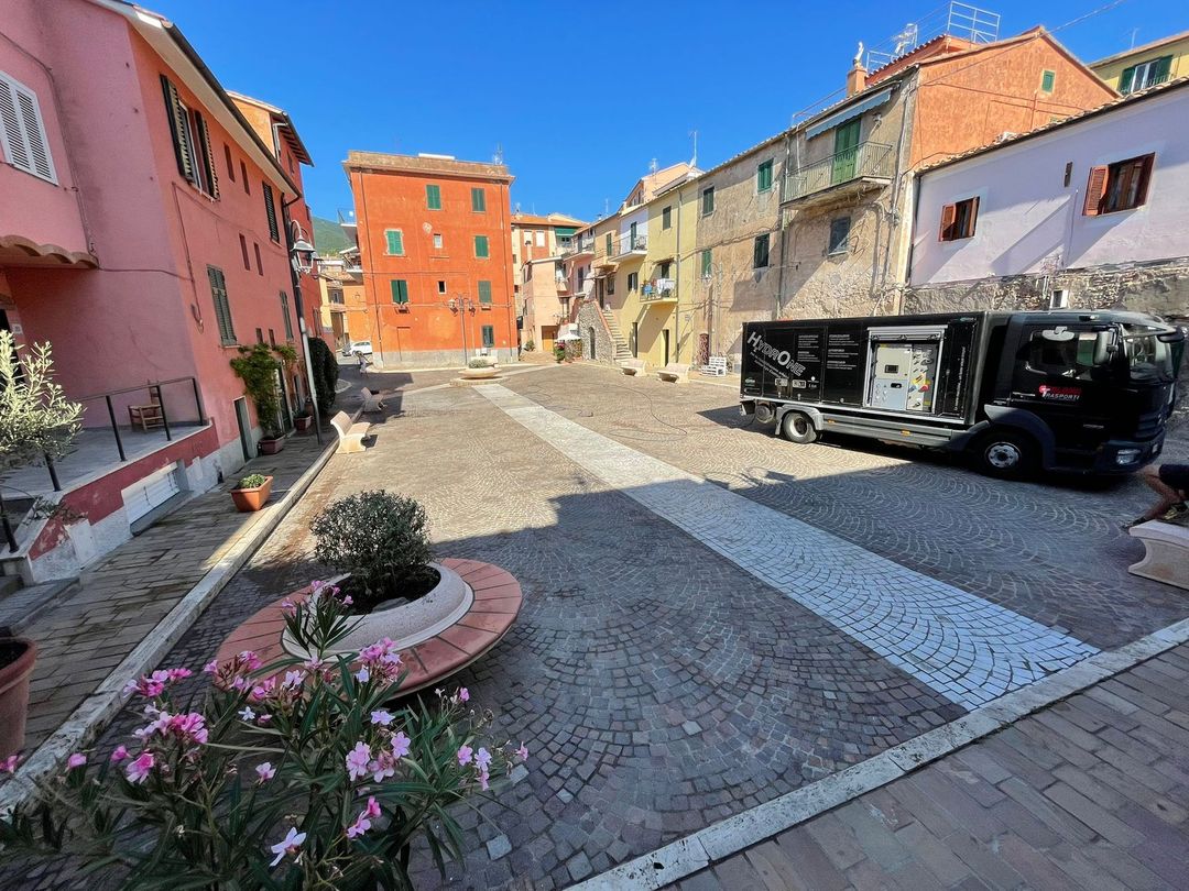 Una soleggiata piazza lastricata in un borgo italiano con edifici colorati e un camion nero parcheggiato.