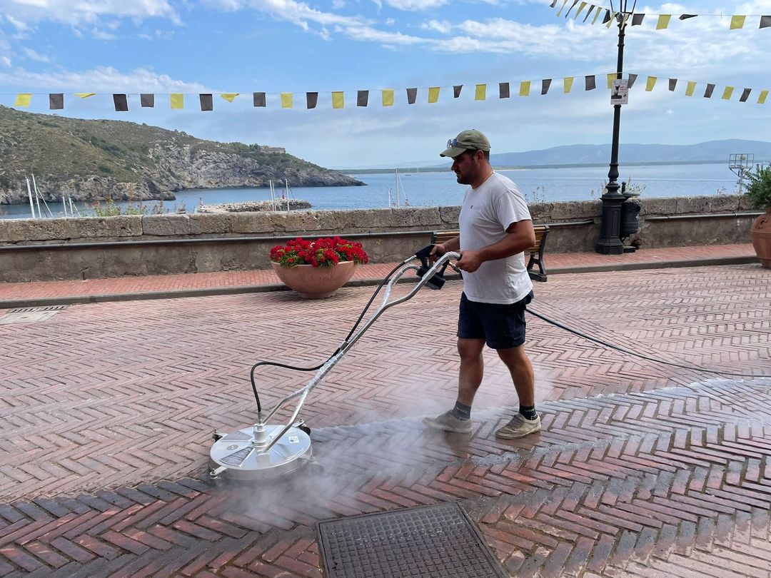 Una persona con una maglietta bianca e pantaloncini scuri usa un detergente per superfici per lavare un sentiero di mattoni a spina di pesce in riva al mare.