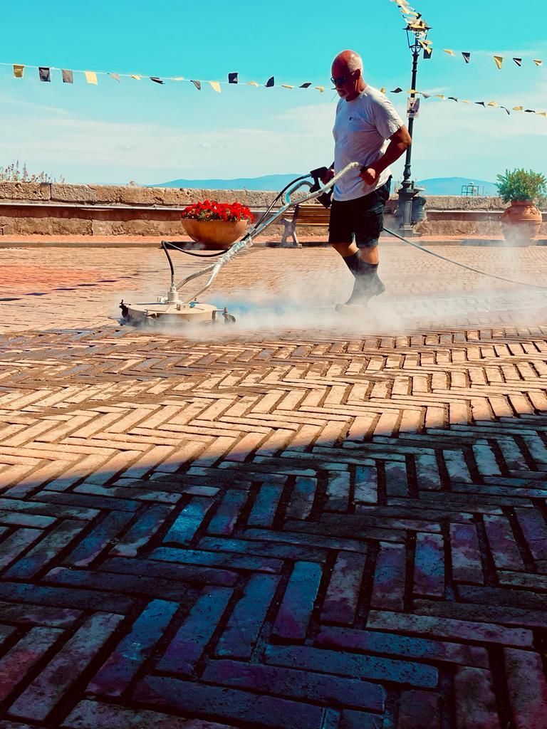 Una persona utilizza un'idropulitrice per pulire un patio in mattoni posati a spina di pesce sotto un cielo limpido e soleggiato.