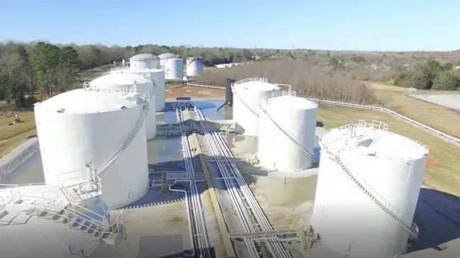 White storage tanks in an industrial area, with connected pipelines and a clear, sunny sky in the background.