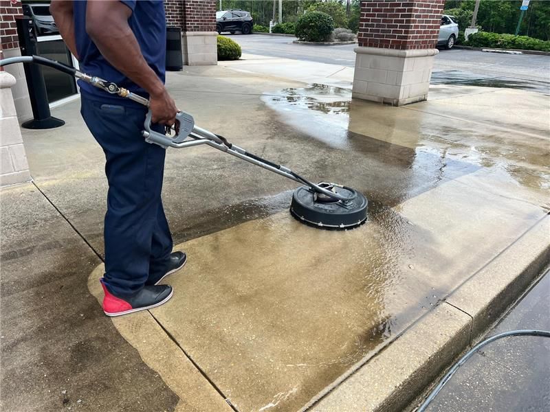 A worker cleaning a concrete sidewalk using a circular pressure washer surface cleaner.