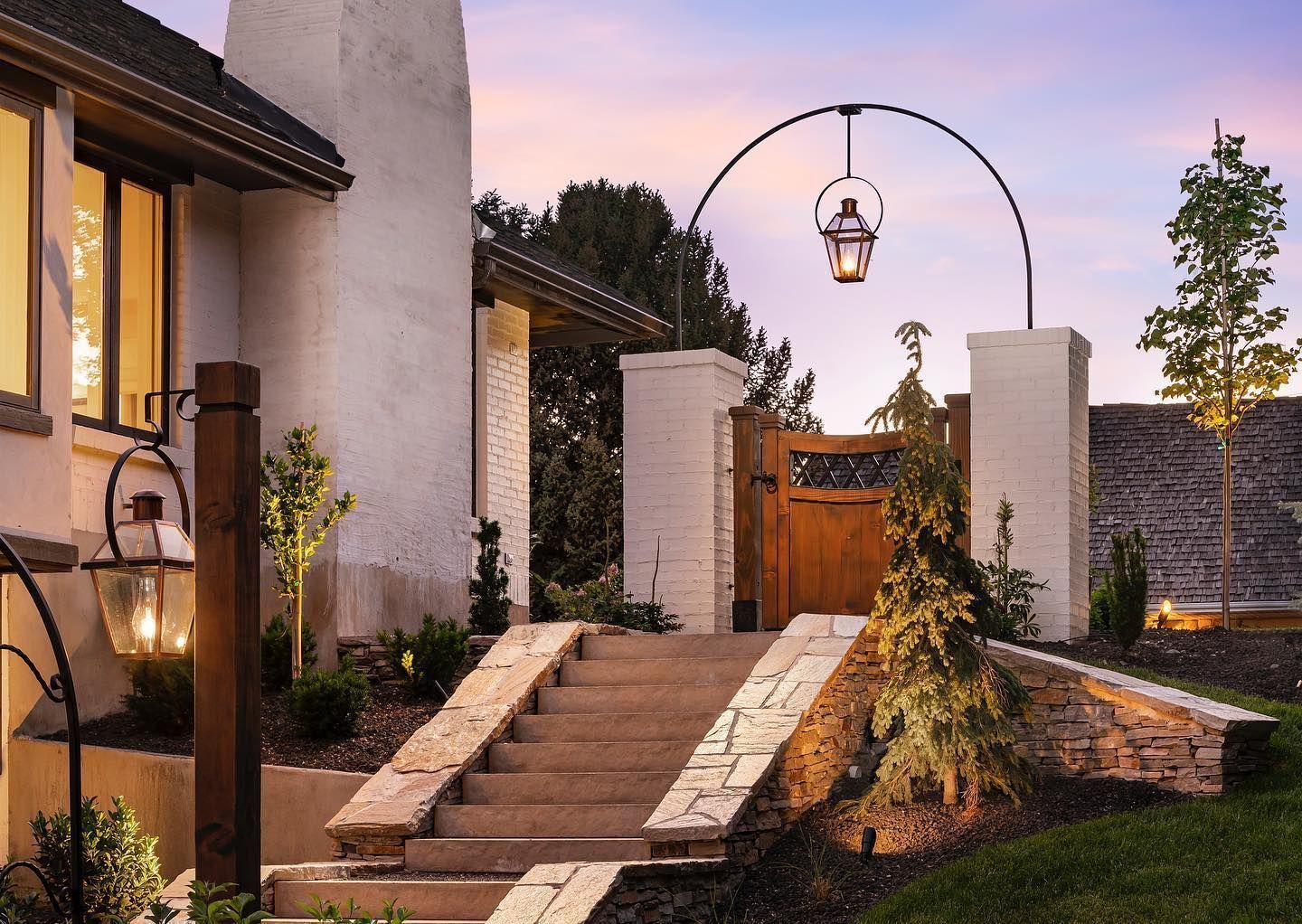Stone steps lead up to a wooden gate framed by white pillars and an arched lantern in a landscaped yard at dusk.