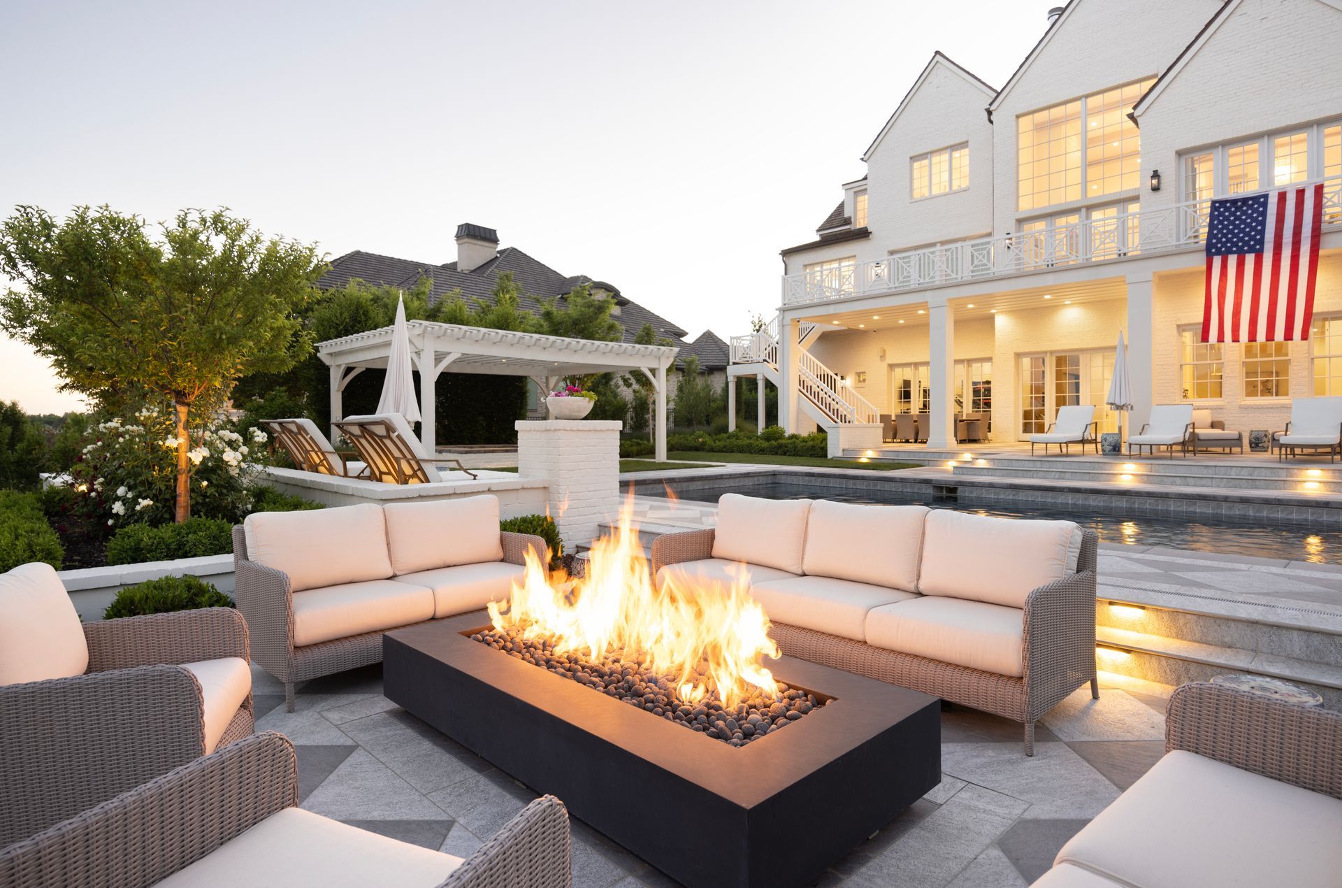 A tiered stone patio with a water feature, lounge chair, gas fire, and landscaped flower pots at dusk.