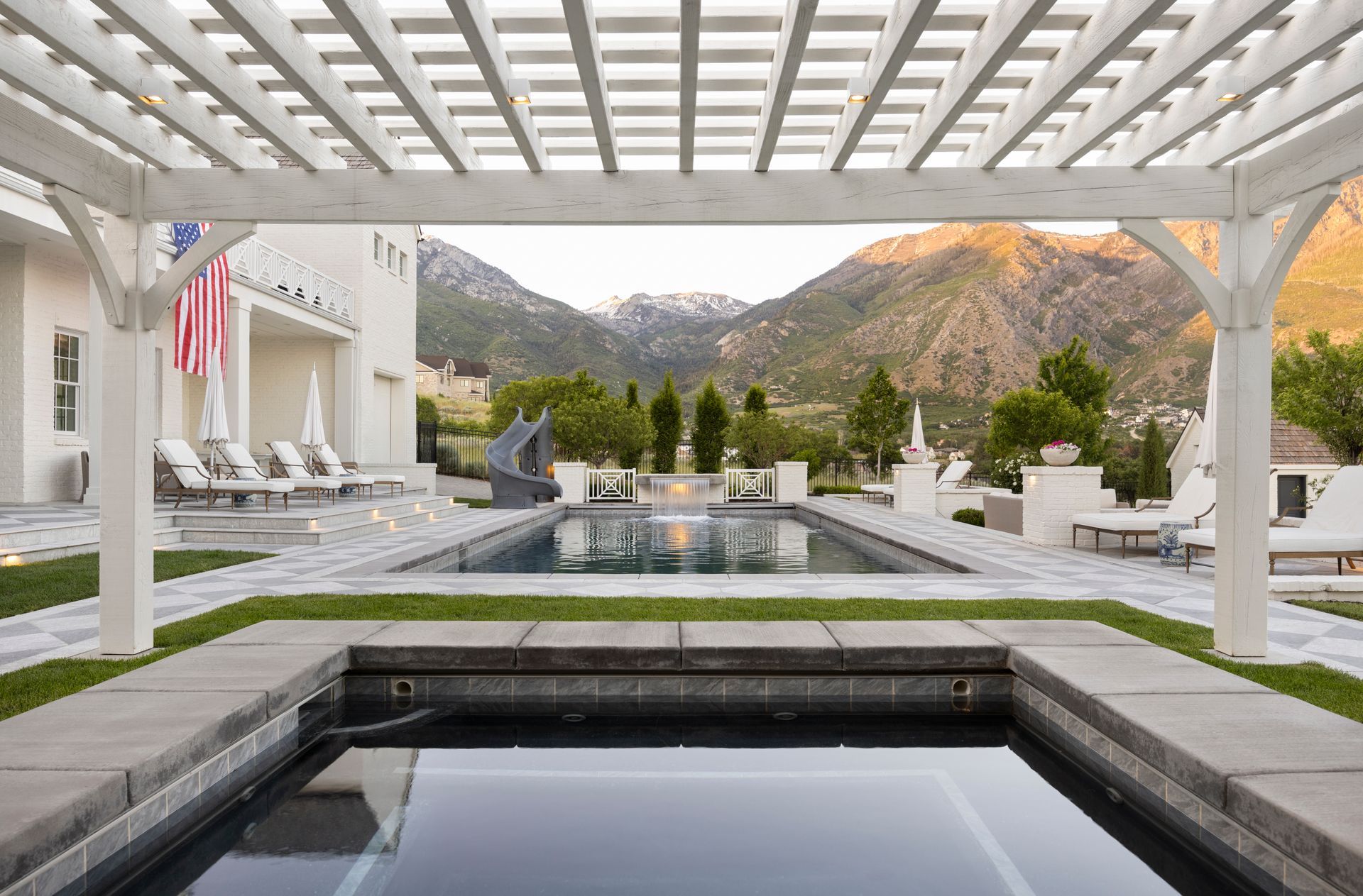 A stone patio with a rectangular swimming pool, a water feature, a waterslide tunnel, and tiered landscaping.