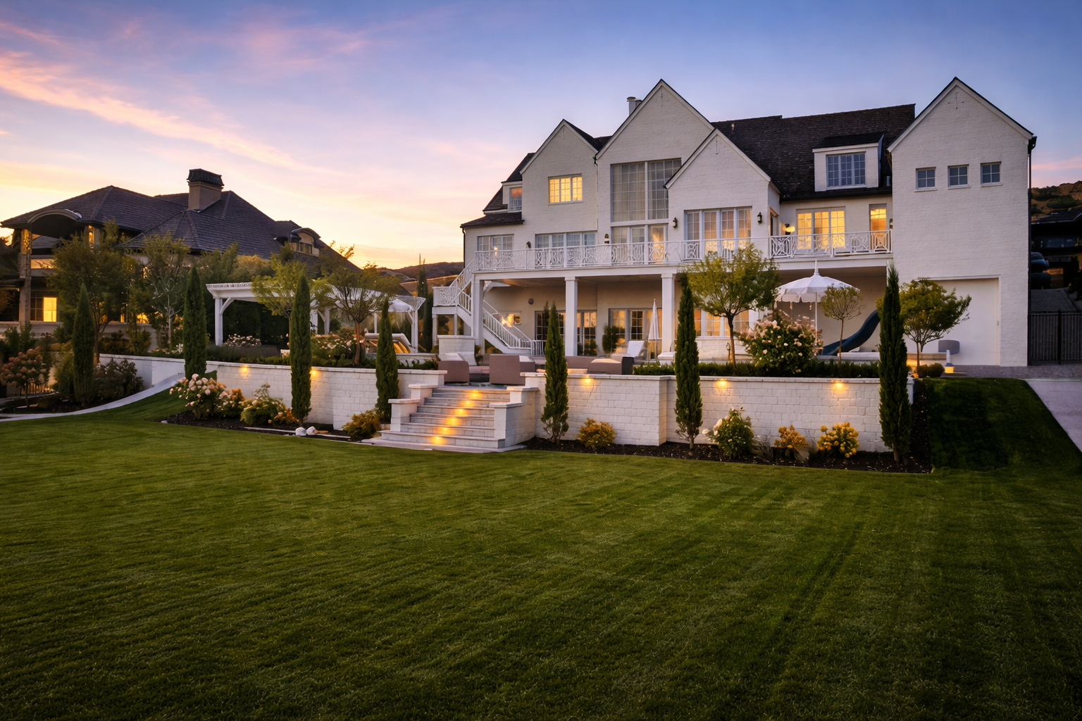 Large, multi-story white house with a manicured lawn, stone terrace, and glowing landscape lighting at dusk.