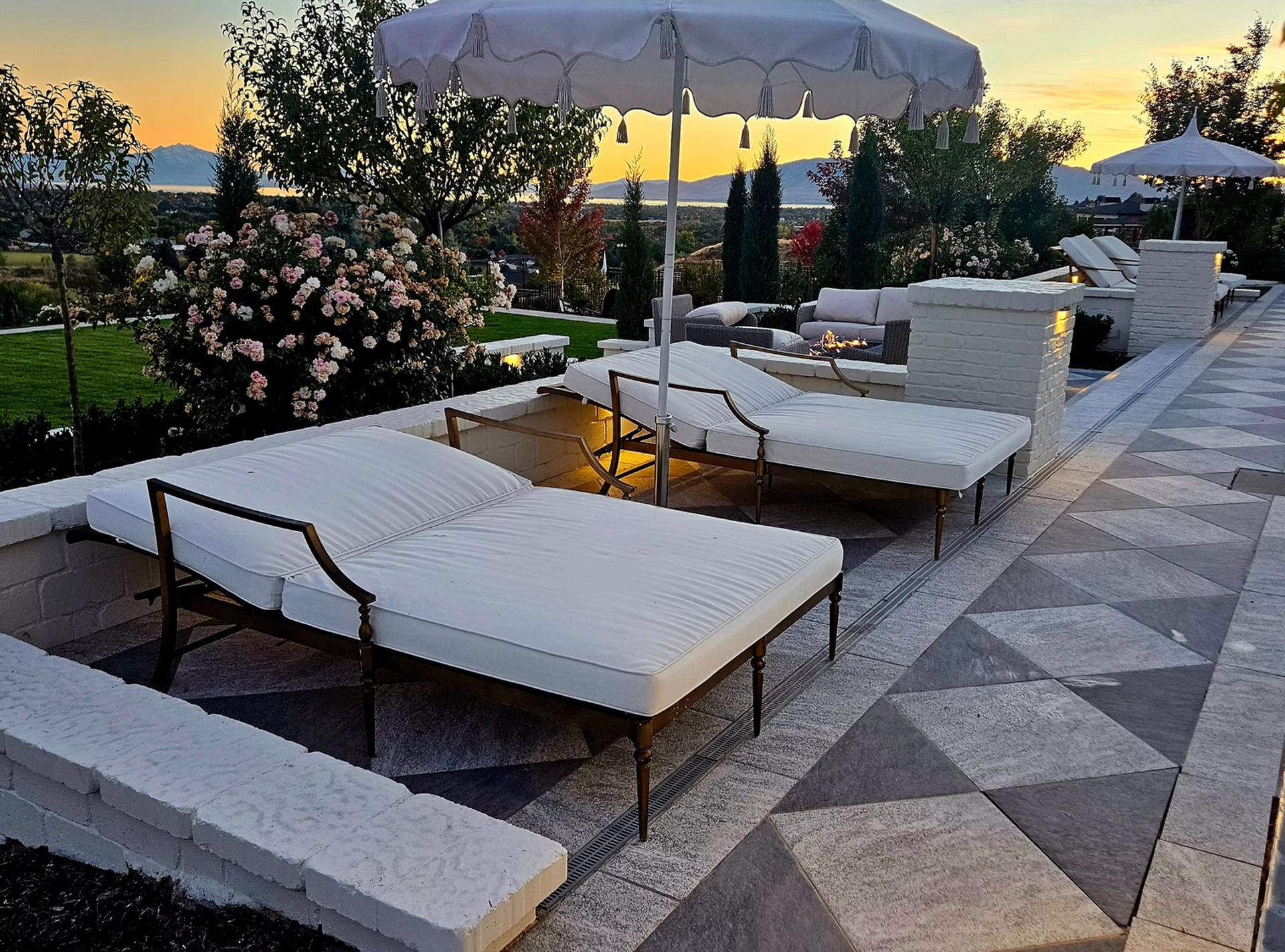 Two white lounge chairs under a patio umbrella on a patterned stone patio at sunset, overlooking a garden and hills.