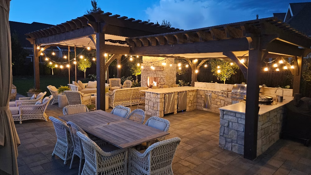 An outdoor patio at dusk featuring a wooden pergola, dining set, stone fireplace, and built-in kitchen with string lights.