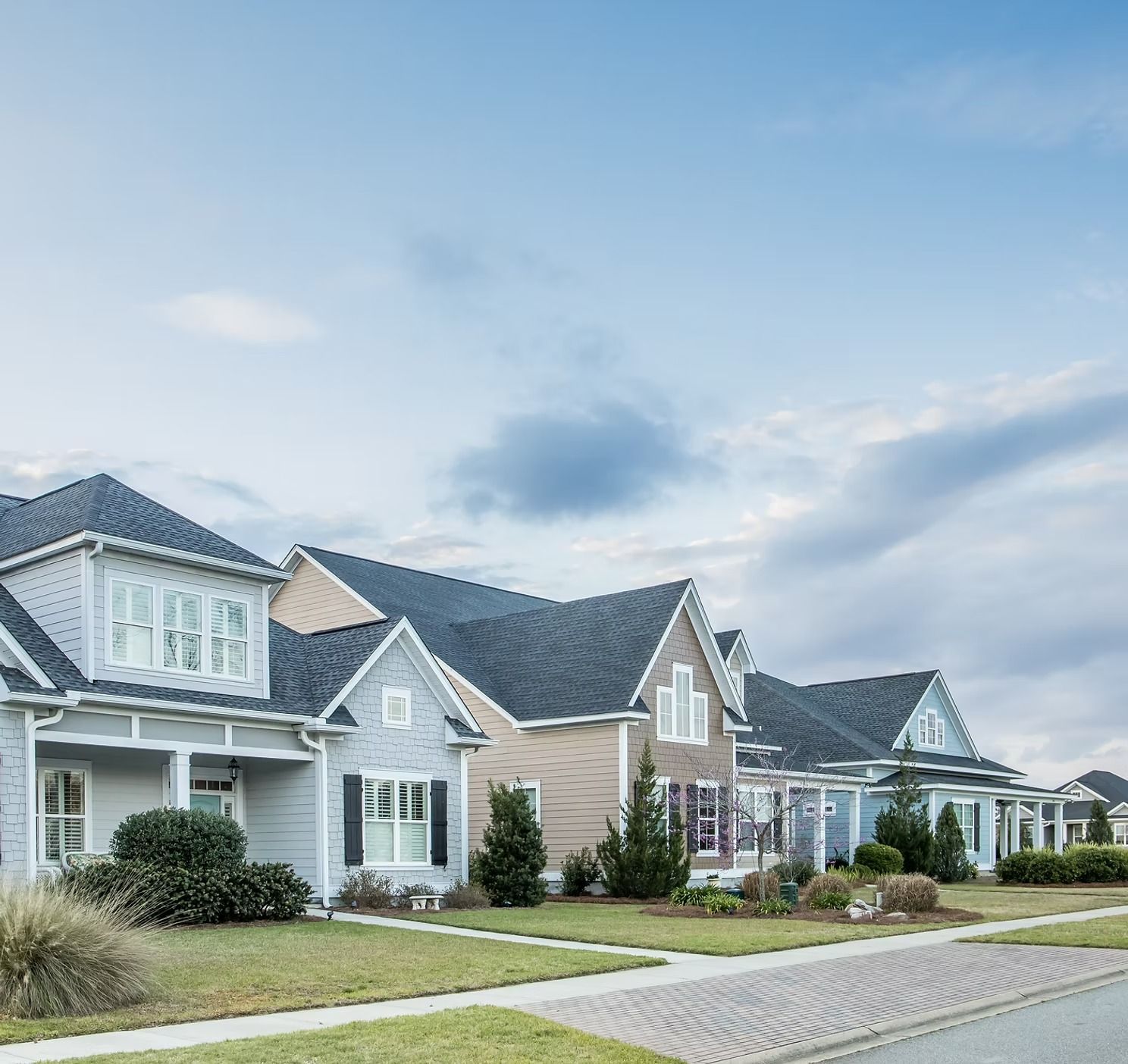 A row of suburban houses with light-colored siding and dark shingled roofs under a blue, cloudy sky.