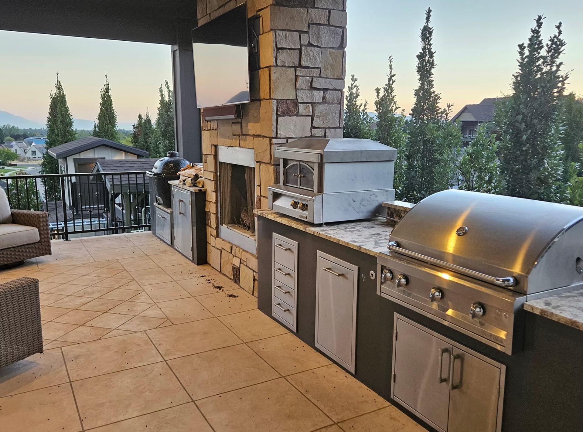 Outdoor kitchen featuring a stone fireplace, stainless steel pizza oven, grill, and cabinets on a tiled patio at sunset.