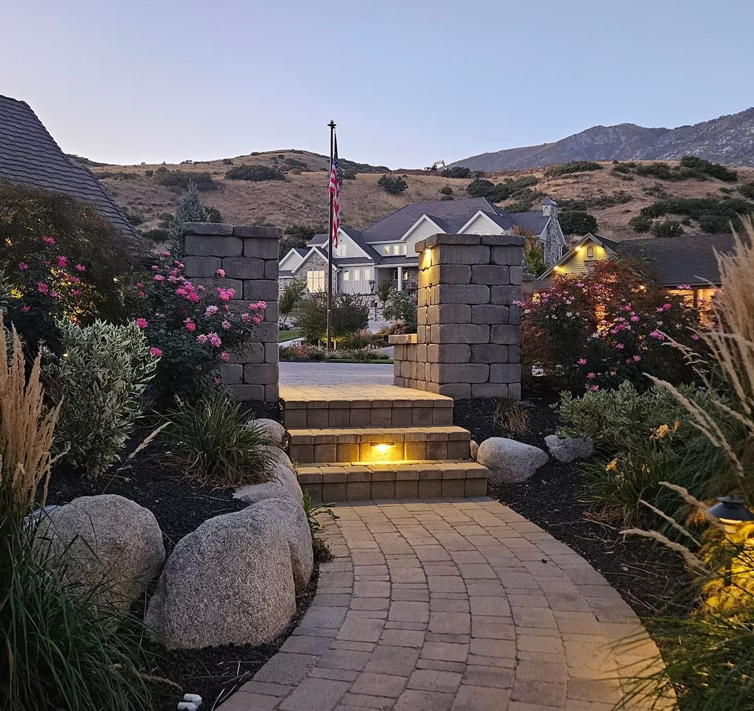 A paved garden path leads to stone steps with integrated lighting, flanked by boulders, rose bushes, and a flagpole.