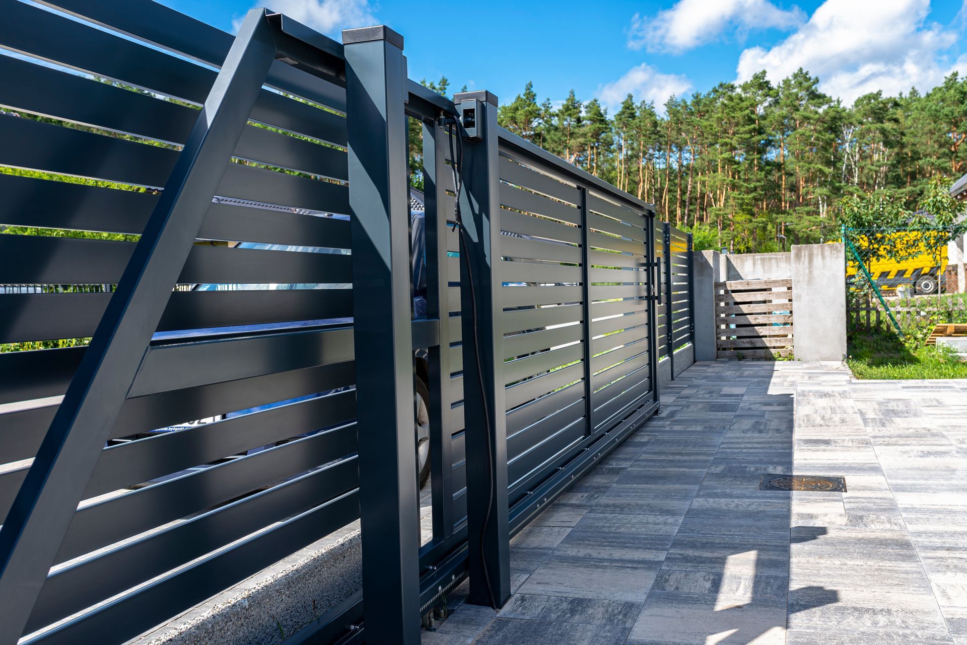 Black sliding gate, partially open, on a paved driveway. Slatted design with a background of trees.