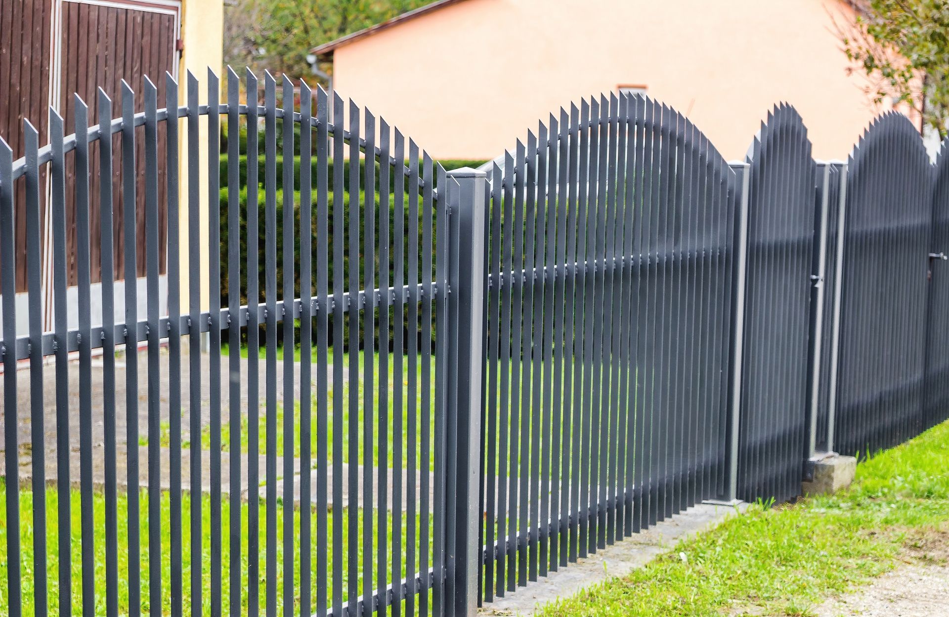 Black metal fence in front of a green lawn, with a house and wooden building visible in the background.
