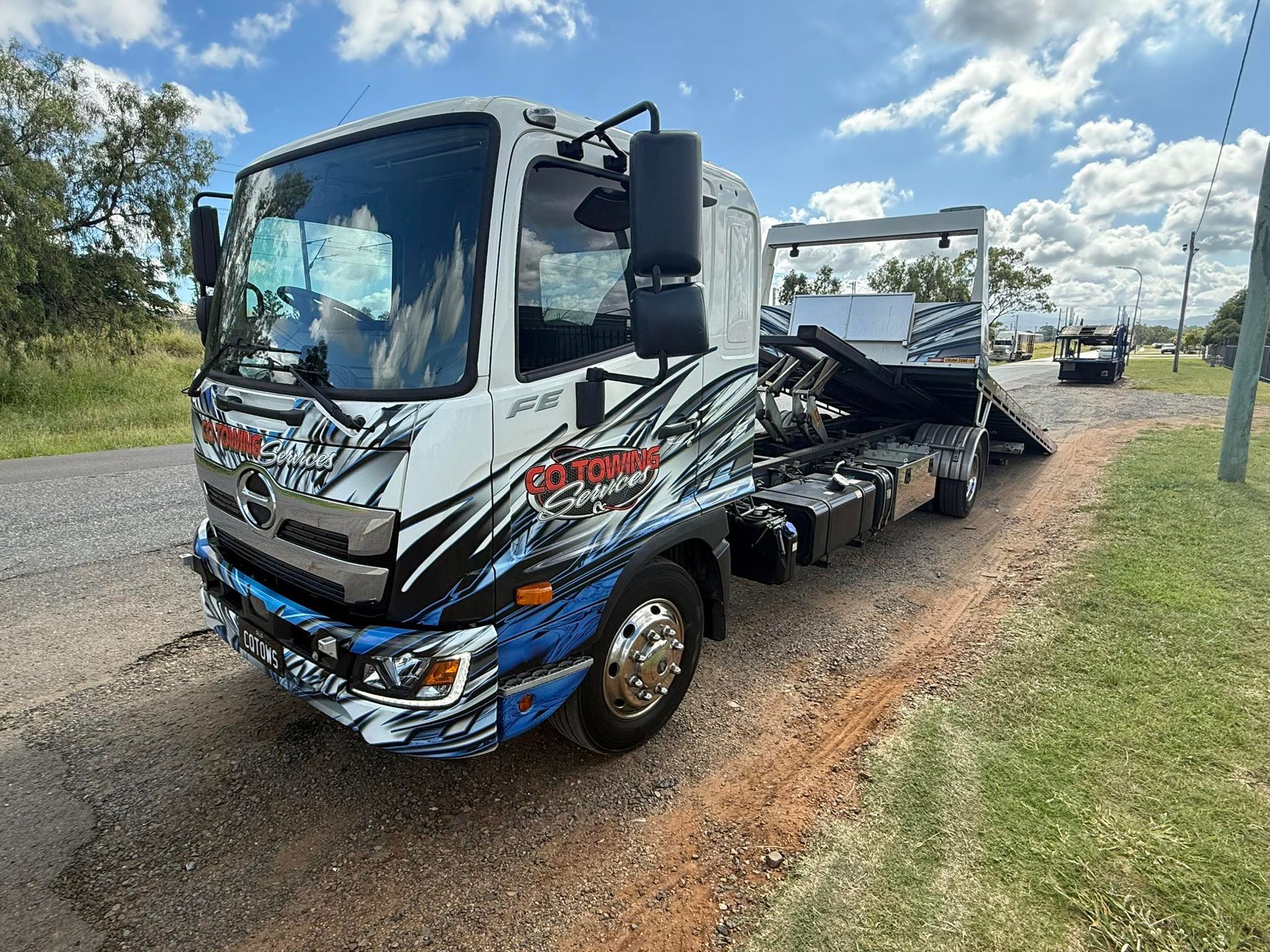 White and blue tow truck parked on the side of a rural road under a partly cloudy sky.