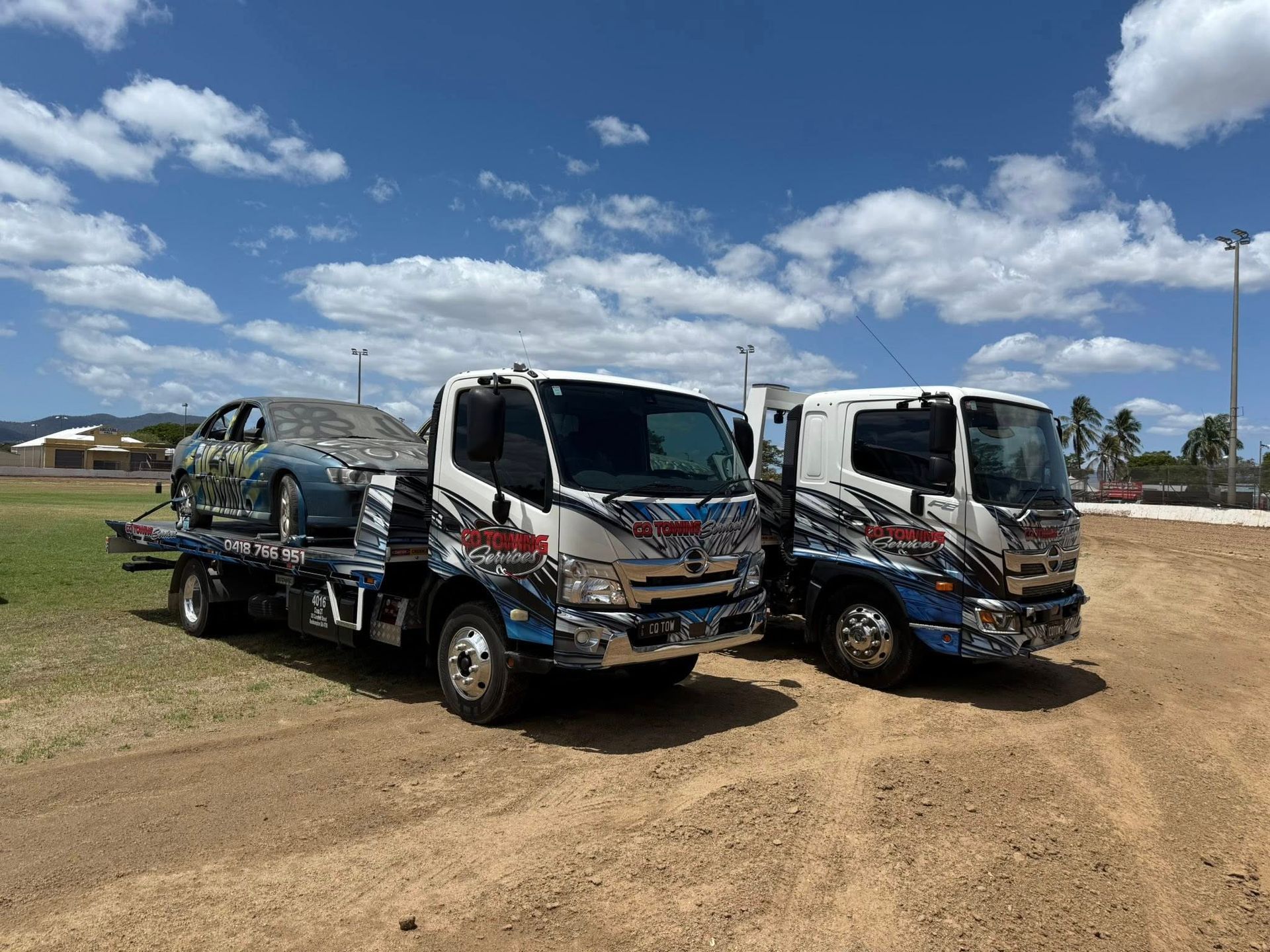 Two tow trucks, one hauling a car, parked on dirt under a blue sky.