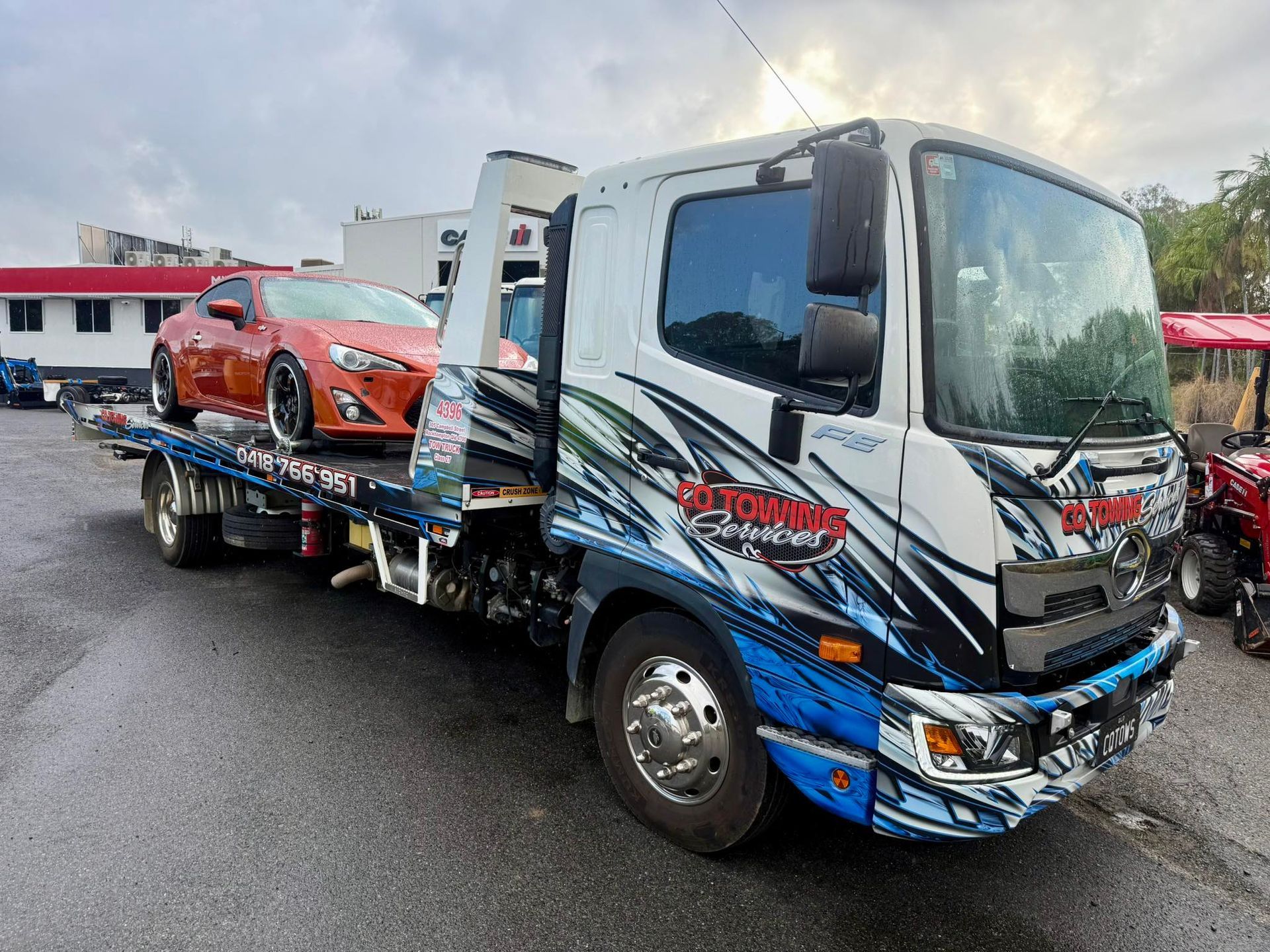 Tow truck with orange car on its flatbed in a parking lot. The truck has blue and white graphics.