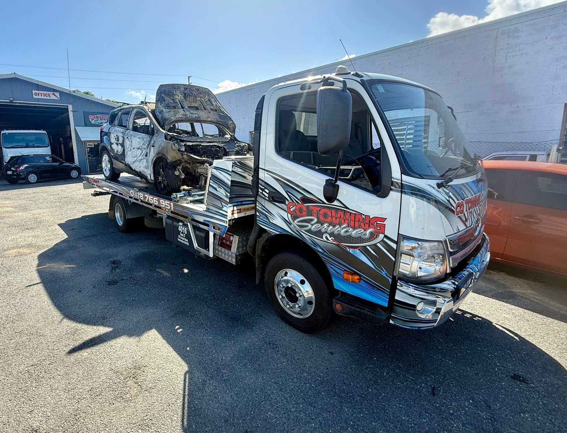 Tow truck carrying a severely damaged, burnt-out car in a sunny outdoor setting.
